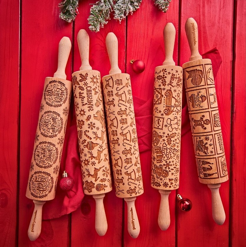 Five Pastrymade Christmas rolling pins with engraved patterns on a red wooden table